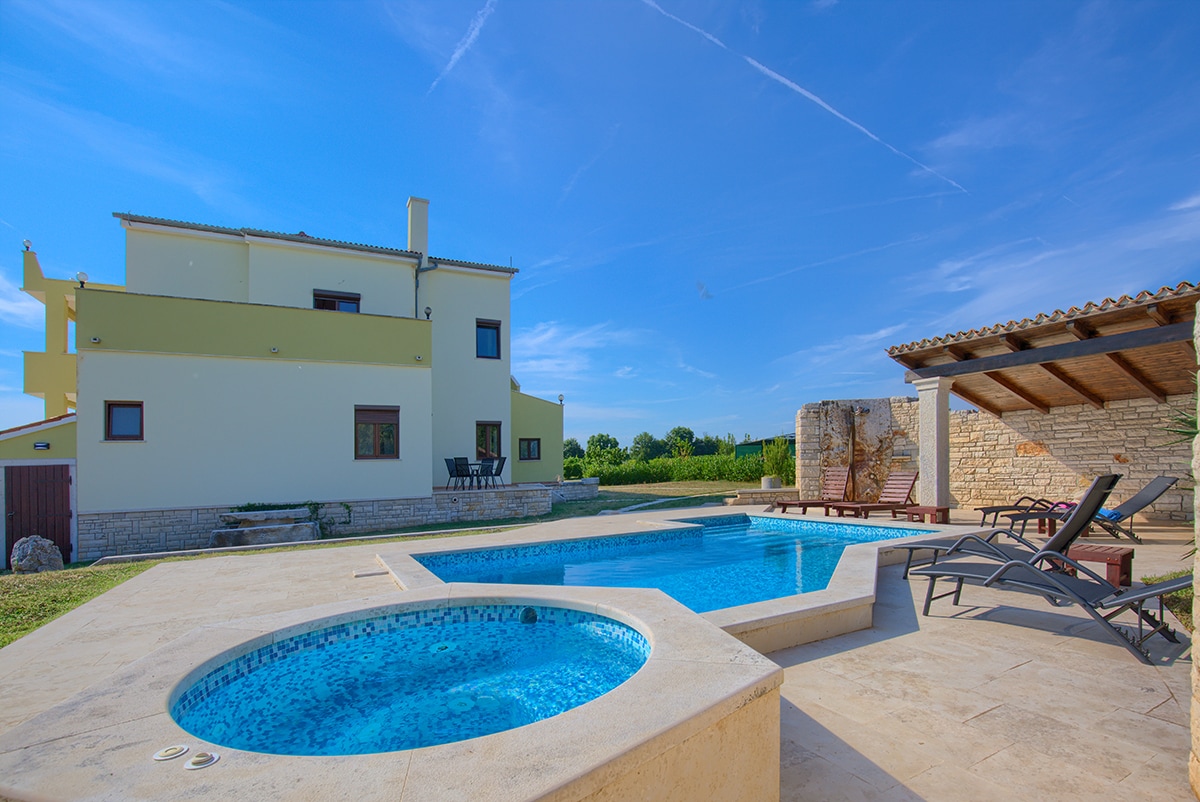 A clear blue sky enhances the serene outdoor area, featuring a swimming pool and a hot tub. Sun loungers are arranged alongside the pool, while the two-story family house is visible in the background, surrounded by green landscapes.