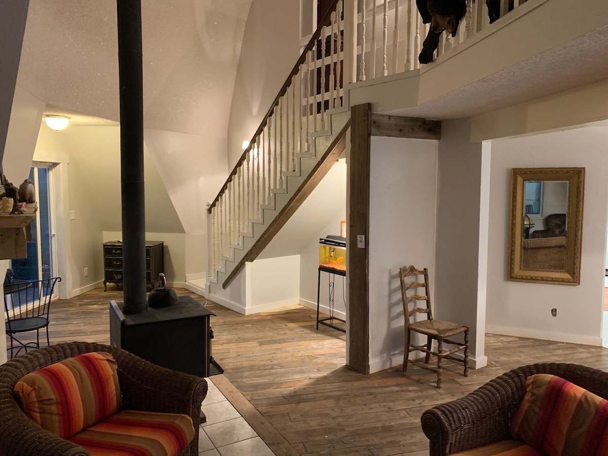 An open-concept living area features a wooden staircase leading to the upper level. Two cozy armchairs with bright cushions are positioned near a metal stove. A large mirror reflects the light from the room, while a television is visible in the background.