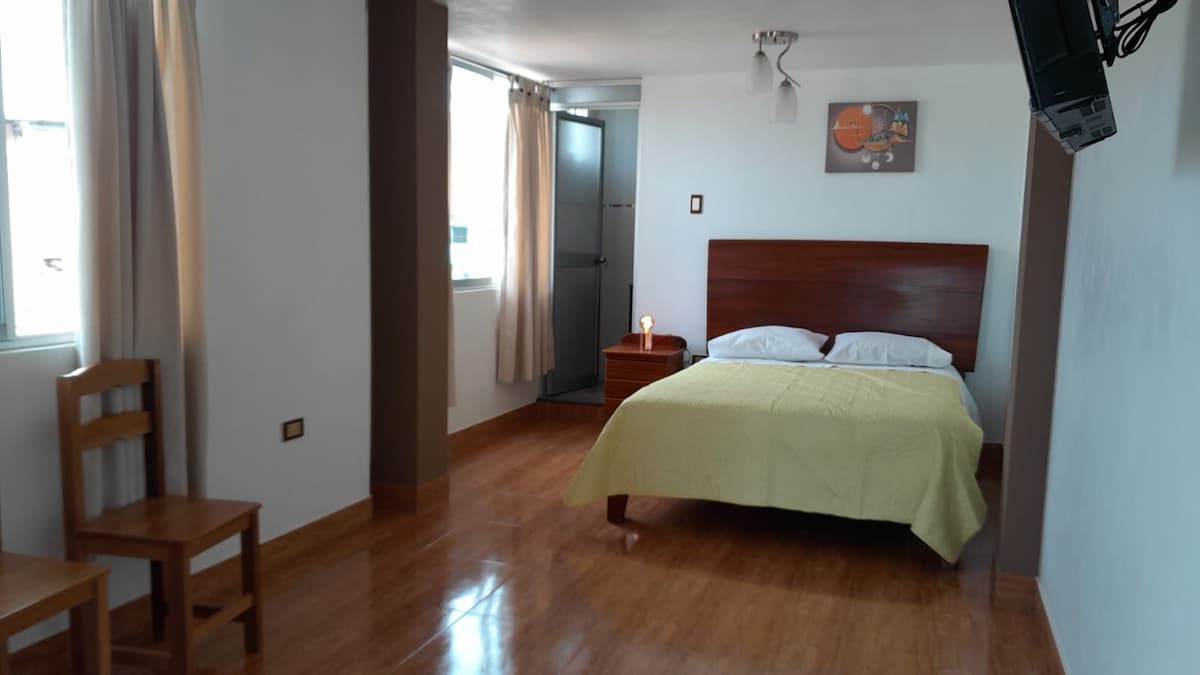 The room features a spacious bed with a light-colored blanket, complemented by a wooden nightstand with a lamp. A simple chair is positioned nearby. Natural light pours in through two windows, with light curtains framing the view. A flat-screen television hangs on the wall.