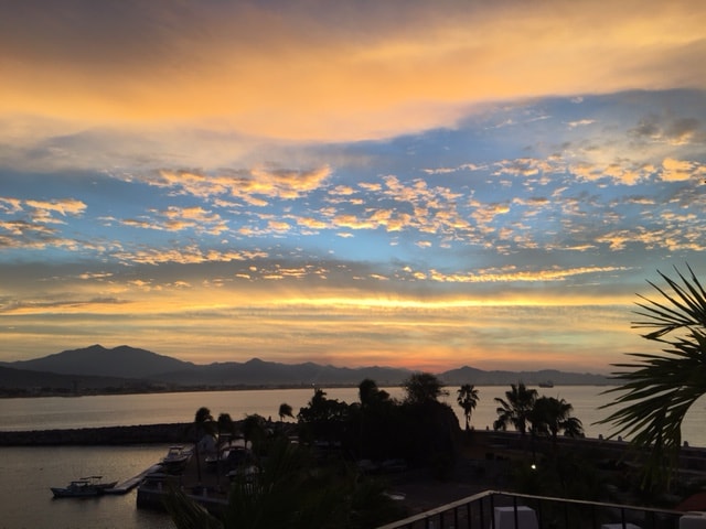 The image captures a panoramic view of a sunset over the bay, with soft clouds painted in shades of orange, blue, and yellow. Silhouettes of mountains and palm trees are visible along the shoreline, enhancing the serene and tranquil ambiance.