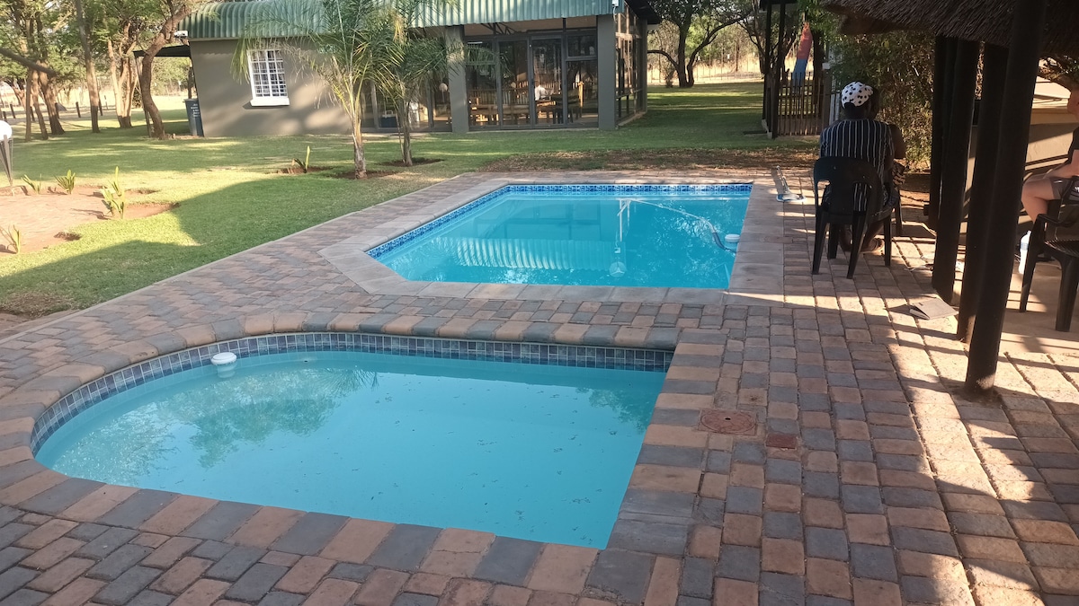 Two swimming pools are framed by stone pavers, one larger and rectangular, and the other smaller and oval-shaped. Lush green grass surrounds the area, with palm trees and a distant lodge visible in the background, creating a serene outdoor space.
