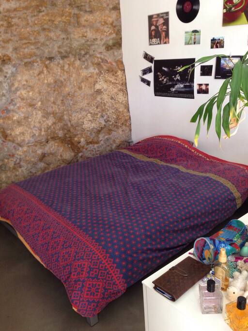 Grand double lit presque carré avec duvet et coussins. Petit buffet de séparation avec bambous pour délimiter le salon et le lit. Mur en pierre.