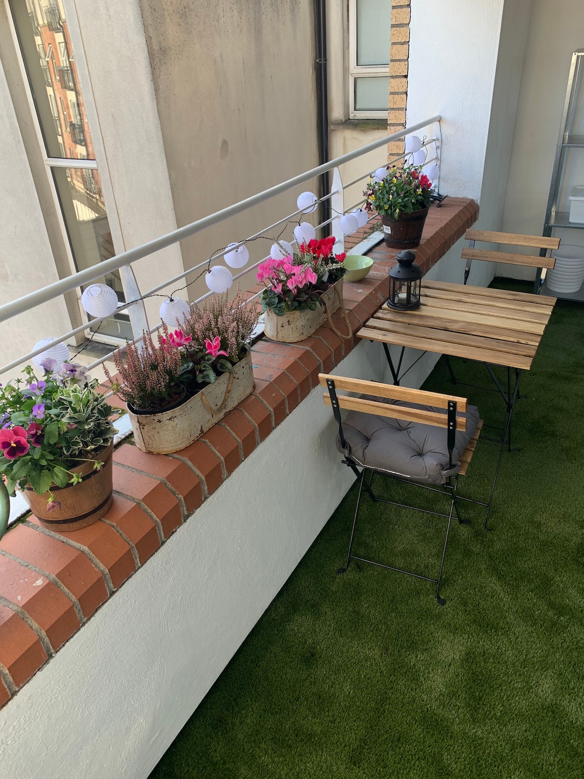 A balcony is featured with vibrant potted plants arranged along a brick railing. Two outdoor chairs are set at a small wooden table, inviting a quiet moment outside. Soft green flooring complements the space, while decorative lights enhance the charming atmosphere.