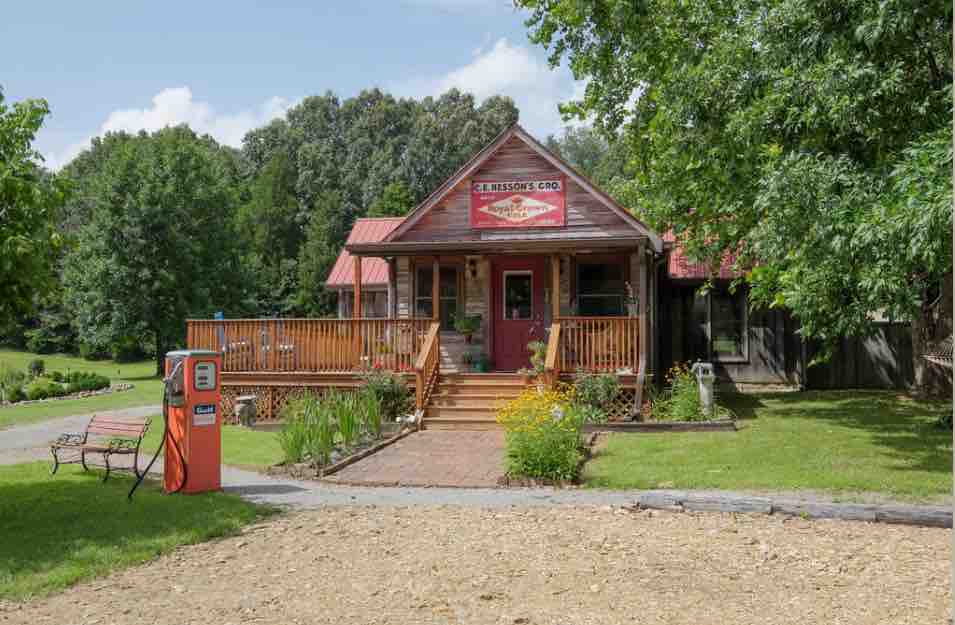 A quaint, single-story house with a welcoming porch is accented by a red metal roof and vibrant signage. Lush greenery surrounds the property, with a gravel path leading to the entrance. A vintage orange pump adds a nostalgic touch to the landscape.
