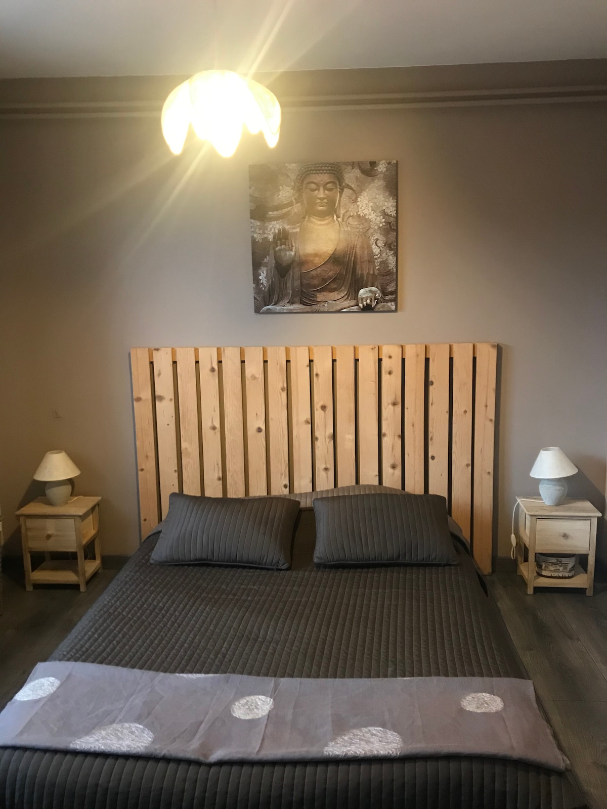 A spacious bedroom features a wooden headboard and a patterned blanket laid over the bed. Two bedside tables with lamps are positioned on each side. A piece of artwork is displayed above the headboard against a neutral wall, complementing the overall serene ambiance.