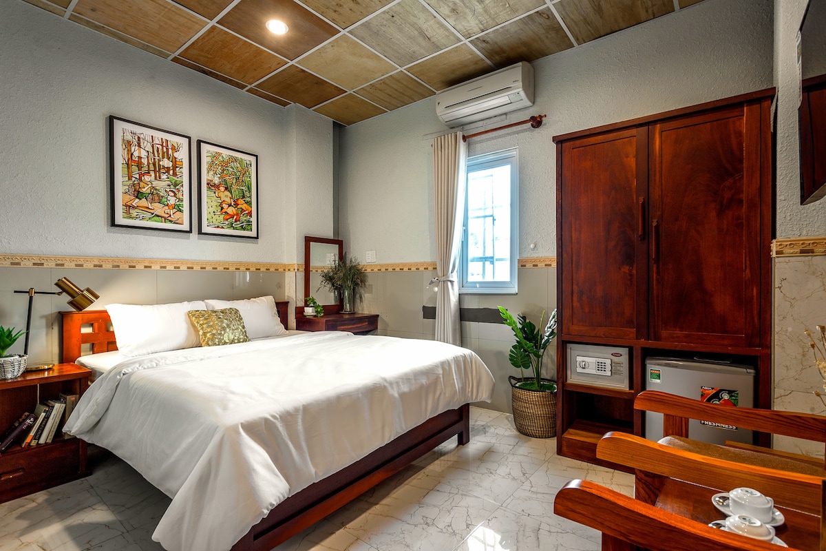 A calming bedroom is presented, featuring a spacious bed with white bedding. Framed artwork adorns the walls, and wooden furniture, including a wardrobe and a small table, enhances the decor. Natural light enters through a window, complemented by a potted plant.