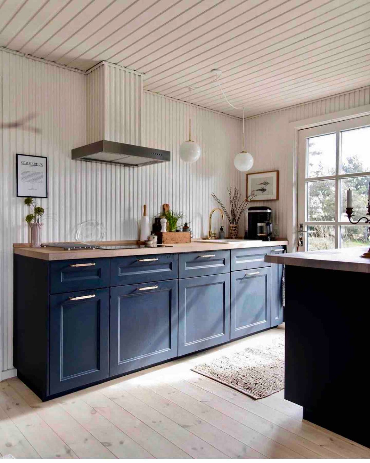A modern kitchen is featured, showcasing navy blue cabinetry with sleek handles. A wooden countertop complements the space, while a minimalist stove hood hangs above. Natural light enters through a window, illuminating the neatly arranged kitchenware and decor, enhancing the warm ambiance.
