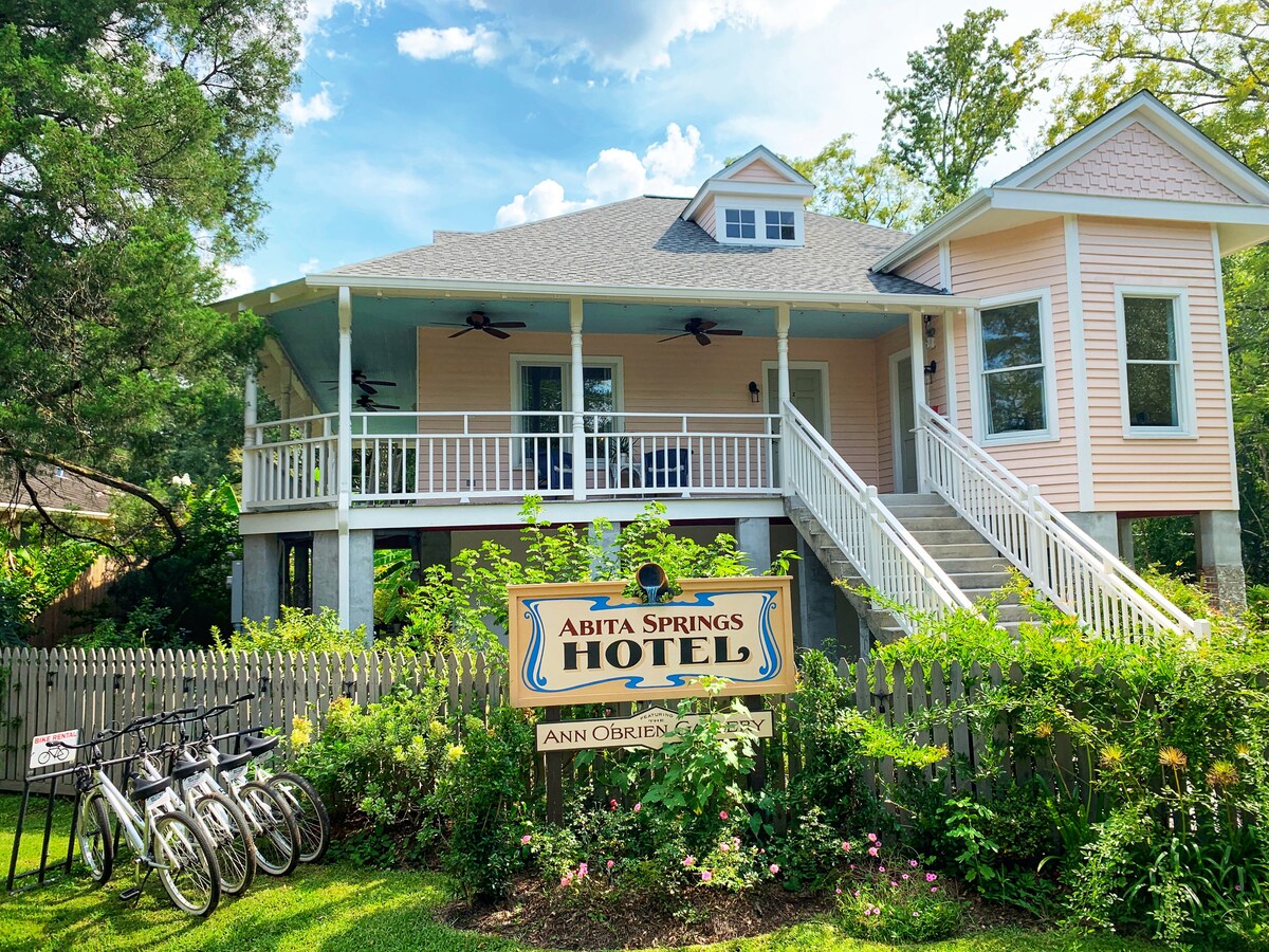 The exterior of a charming pink hotel is displayed, featuring a wraparound porch with ceiling fans and steps leading to the entrance. A sign reading 'Abita Springs Hotel' is prominently featured, while bicycles are parked nearby amidst landscaping with lush green foliage.