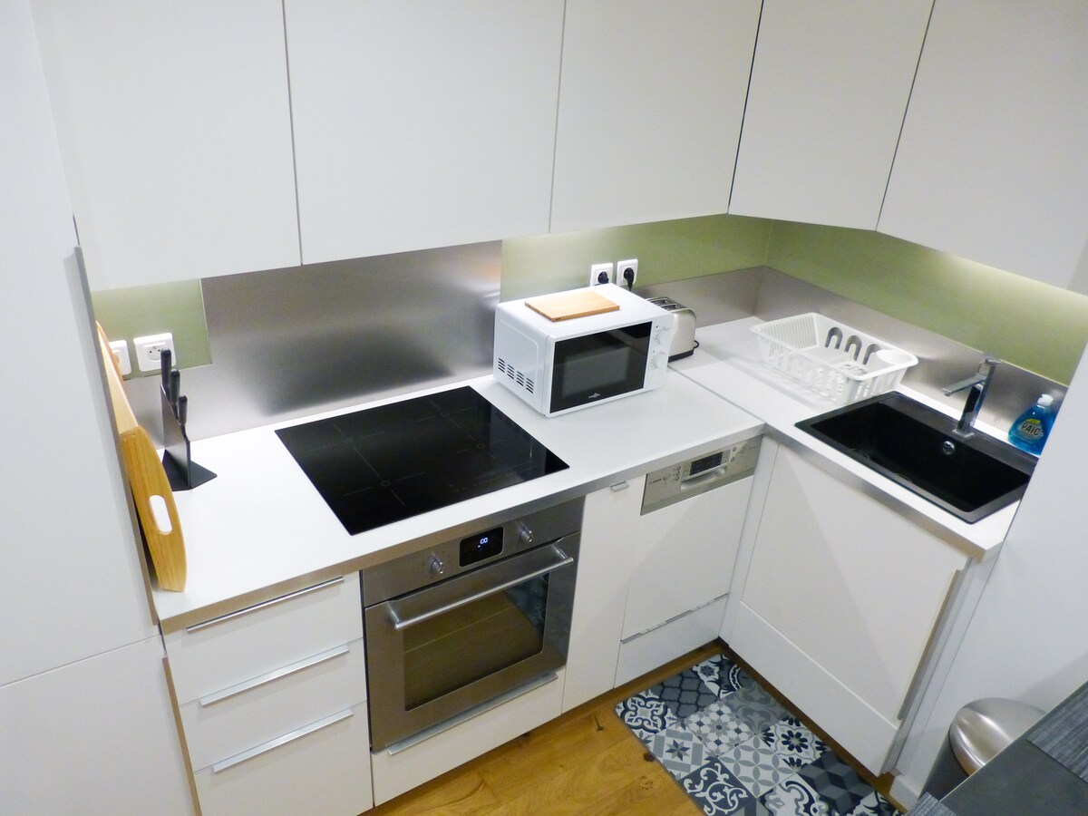 The kitchen is equipped with modern appliances, including an induction cooktop, oven, microwave, and dishwasher. A sleek sink and a white countertop provide ample workspace, while an organized area for utensils and dishware is visible. A patterned rug adds a touch of style on the floor.