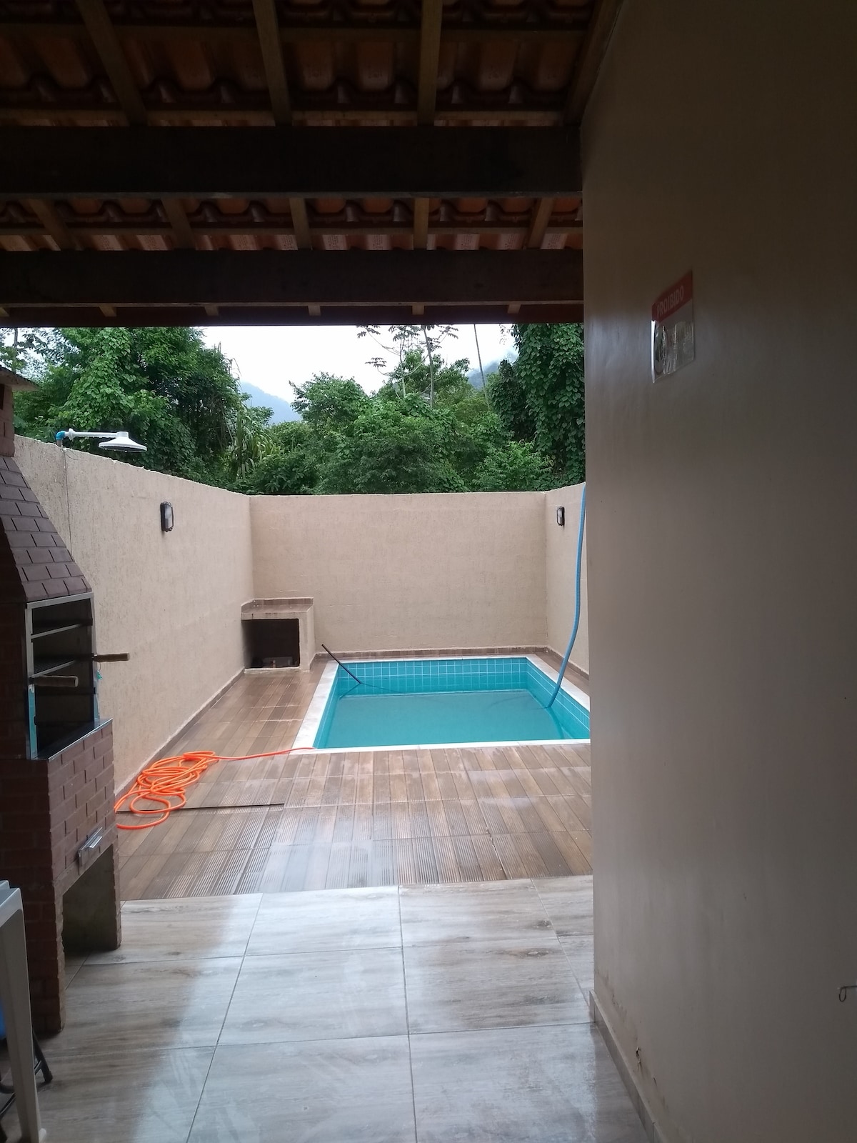 A view from an interior space opens to a private pool area, featuring a rectangular pool set against a textured wall. Wooden decking surrounds the pool, while a brick barbecue grill is visible to the left. Lush greenery is seen beyond the enclosure.