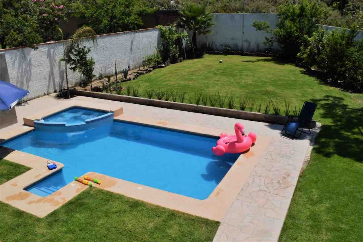 A vibrant swimming pool is surrounded by well-maintained grass and greenery. A hot tub is adjacent to the pool, with a pink inflatable float resting on the surface. Sun loungers are positioned nearby, offering a relaxing space for enjoyment.