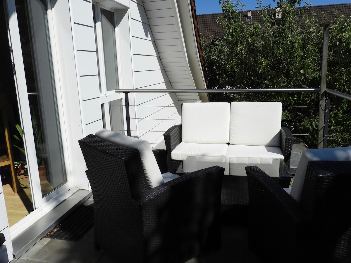 A terrace area features a seating arrangement with a white sofa and two matching armchairs, all set around a small table. The space is shaded by surrounding greenery and is accessible through large glass doors.