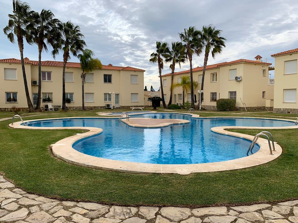 A spacious communal pool is framed by well-maintained greenery and palm trees. The pool features a central island and gentle curves, with a stone path leading around its perimeter, offering a tranquil atmosphere for relaxation.