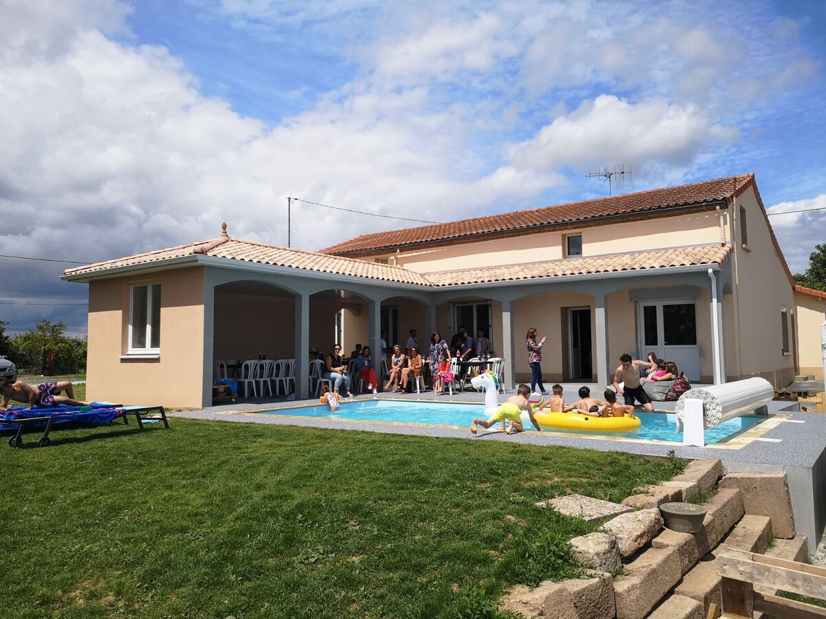 A renovated house features a spacious terrace overlooking a heated pool. Lush green grass surrounds the area, with multiple sun loungers and seating available. Groups of people are engaged in leisure activities, illustrating an inviting atmosphere for family and friends.