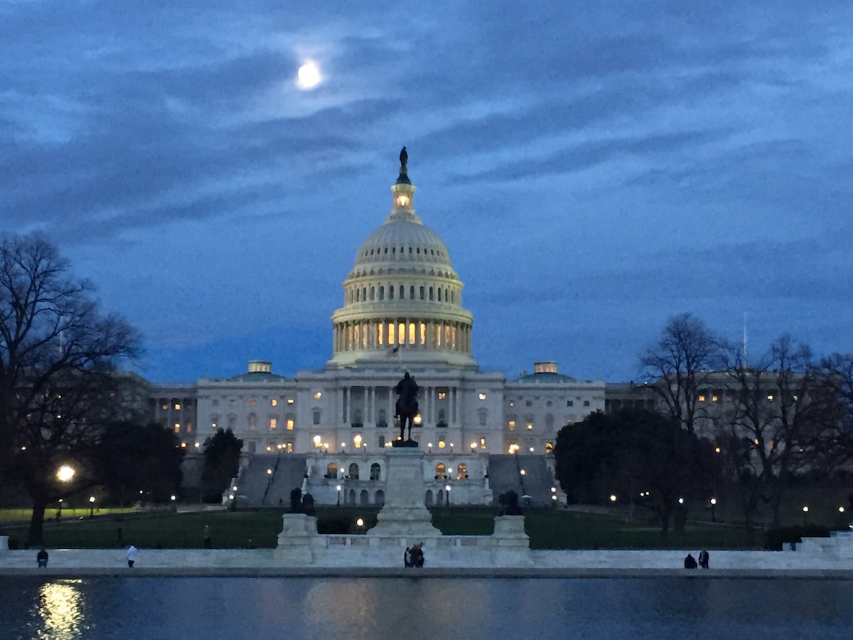 The iconic Capitol building is viewed from a distance at dusk, illuminated by soft lights against a blue sky. A statue stands in the foreground near a reflecting pool, surrounded by trees that are bare of leaves.