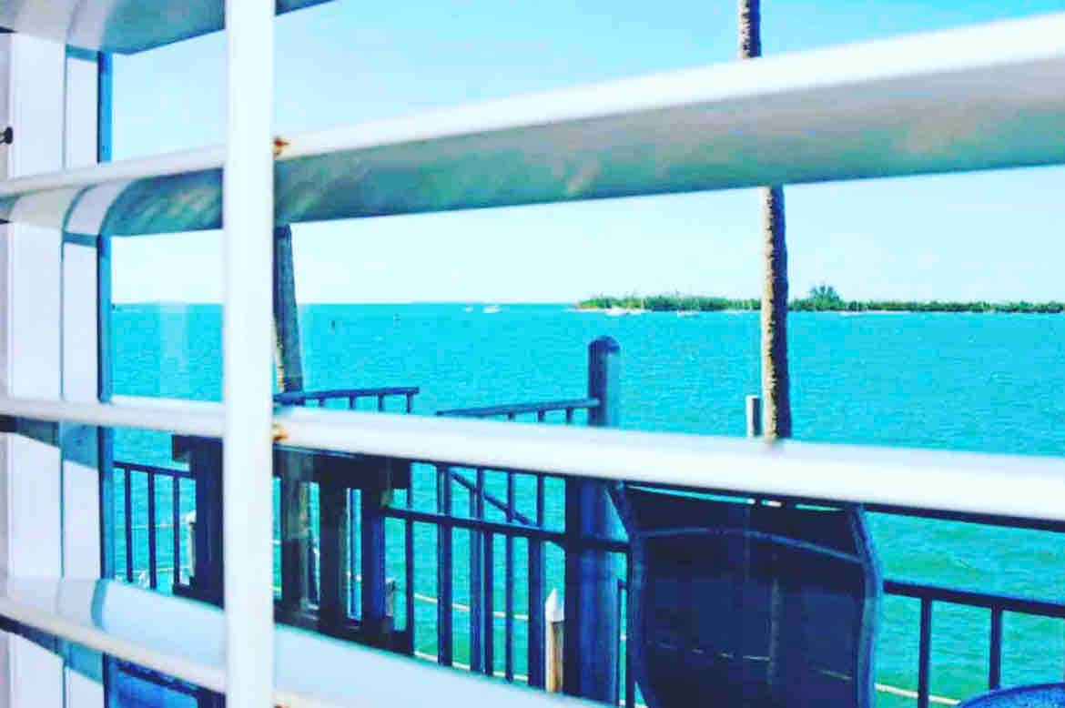 A view of the ocean is framed by horizontal blinds. The turquoise water is visible, reflecting the clear blue sky. In the distance, a small island is seen. A dark chair is positioned on the balcony, complementing the relaxed atmosphere.