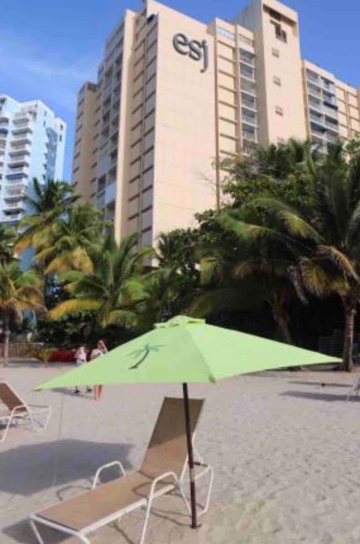 A lime green umbrella provides shade on a sandy beach, surrounded by palm trees. The ESJ Towers building stands in the background, showcasing its distinct architecture. Lounge chairs are positioned nearby, inviting relaxation under the sun.
