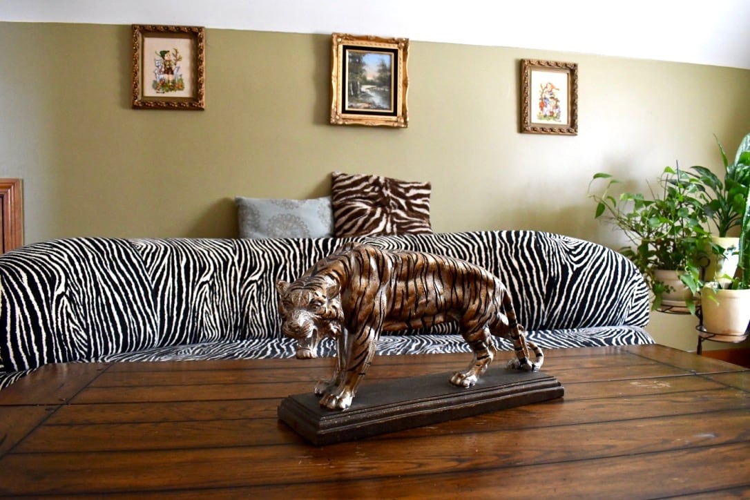 A wooden table is showcased in front of a zebra-striped couch. A decorative tiger sculpture rests on the table's surface. The wall behind features framed artwork, and plants are placed around the room, enhancing the overall decor.