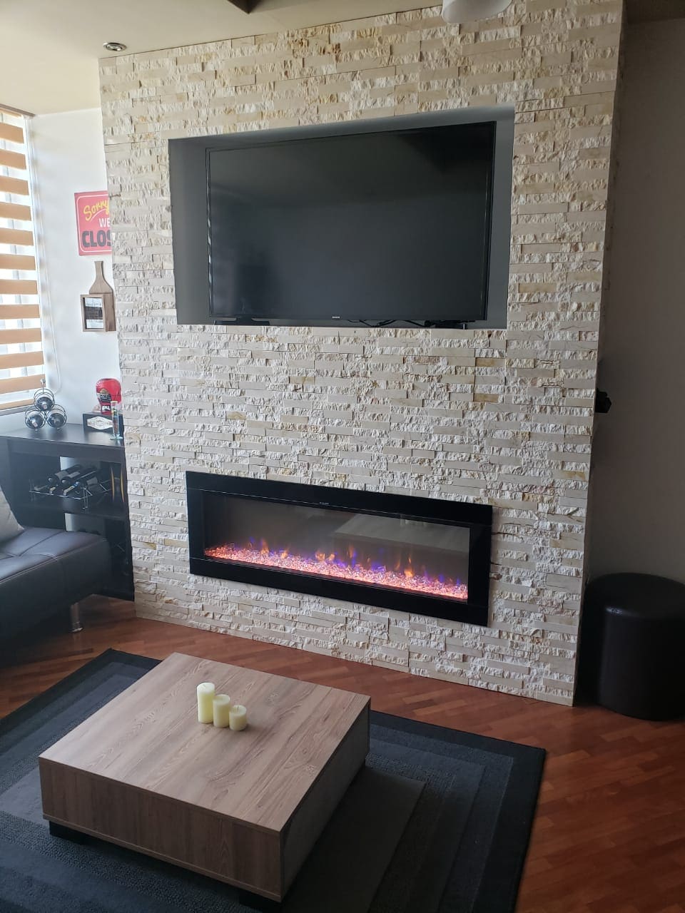A modern living area features a wall adorned with textured stone, highlighting an electric fireplace below a flat-screen television. A low coffee table sits centrally, topped with light-colored candles. Dark furnishings and a rug create a comfortable and balanced aesthetic.