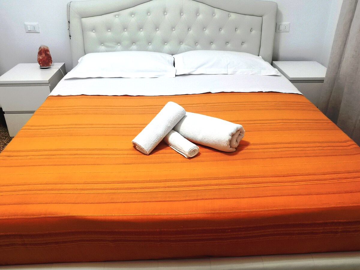 A spacious bed is dressed in a vibrant orange coverlet, complemented by neatly rolled white towels. The tufted headboard adds a touch of elegance, while bedside tables with soft lighting contribute to the room's overall comfort.
