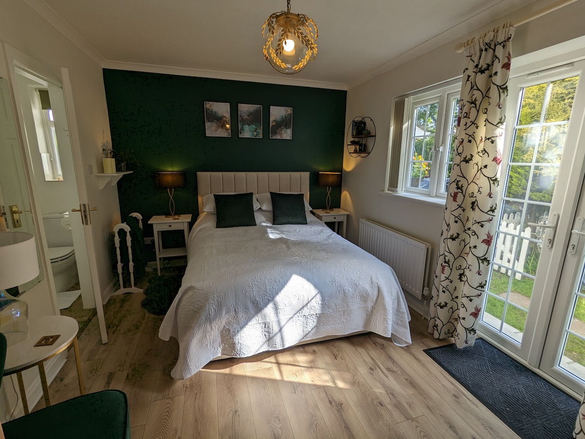 A spacious sleeping area features a large white bed with decorative pillows against a dark green accent wall. Natural light enters through French doors, illuminating the wooden floor. Two bedside lamps and framed artwork complement the serene atmosphere, with floral curtains adding a touch of softness.