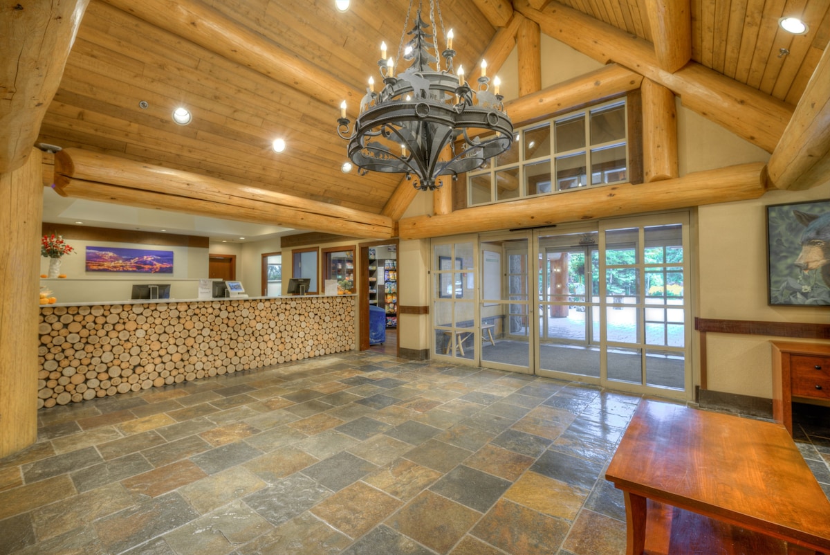 A welcoming lobby is characterized by high, exposed wood beams and a large chandelier. A stone floor complements the natural wood reception area, which features a stacked log wall. Floor-to-ceiling windows provide ample natural light, revealing a view of the outside landscape.