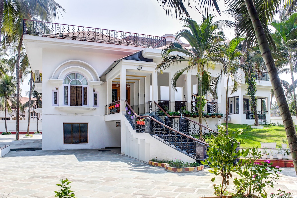 The villa showcases a modern architectural style with a spacious terrace and multiple palm trees surrounding the entrance. An elegant staircase leads up to the covered porch, where decorative floral accents add a touch of color to the exterior.