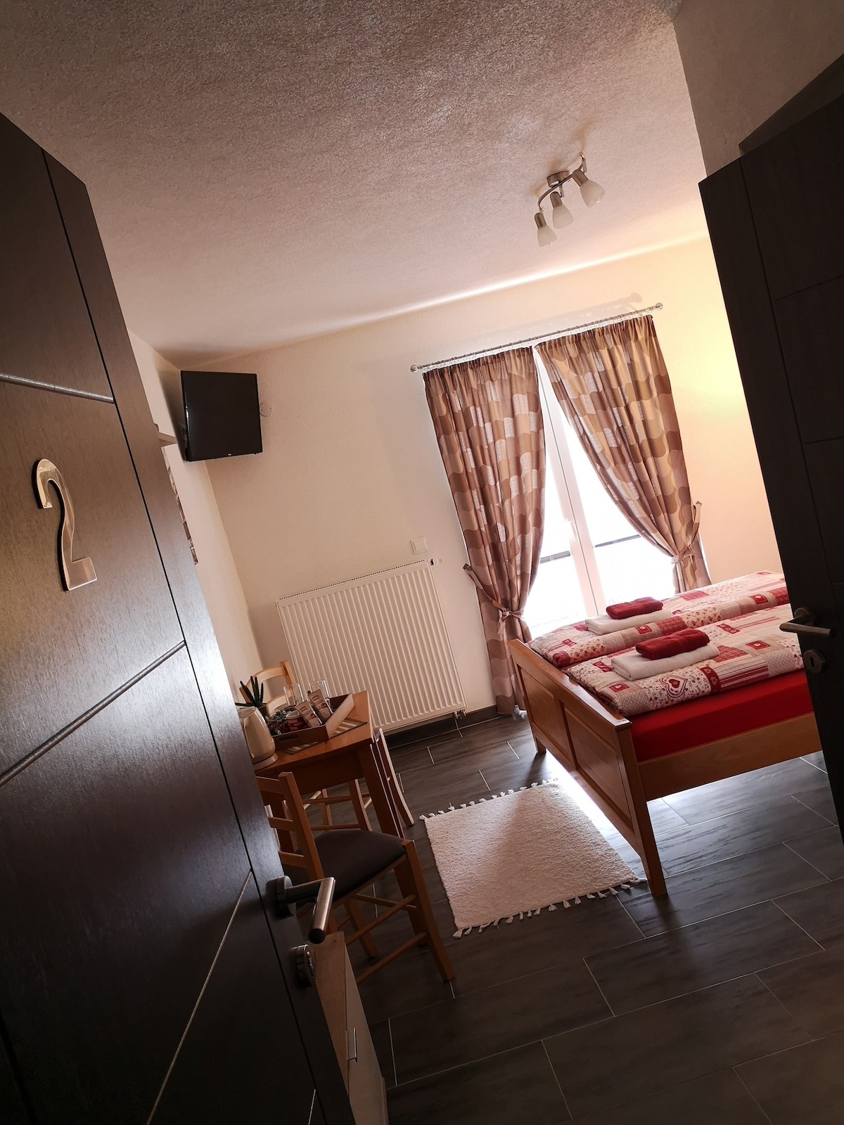 A cozy room is visible through an open door, featuring a red and white bed with decorative pillows. A small wooden table and chairs are positioned near a window dressed in patterned curtains. A flat-screen TV is mounted on the wall, adding convenience.