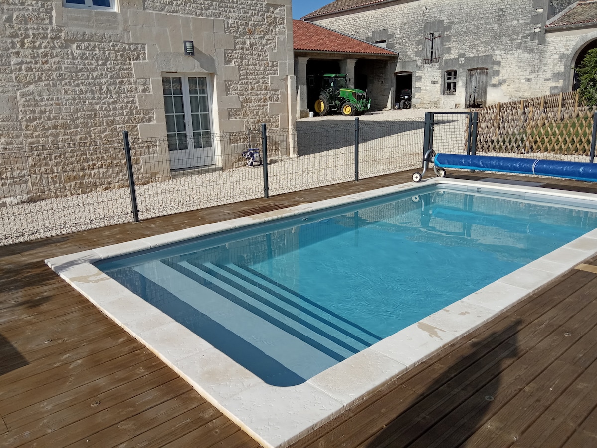 A clear salt-water pool is set against a backdrop of rustic stone buildings. Wooden decking surrounds the pool, enhancing the tranquil setting. A safety fence provides an enclosed area, while a solar cover roller is positioned beside the pool, ensuring convenience for guests.