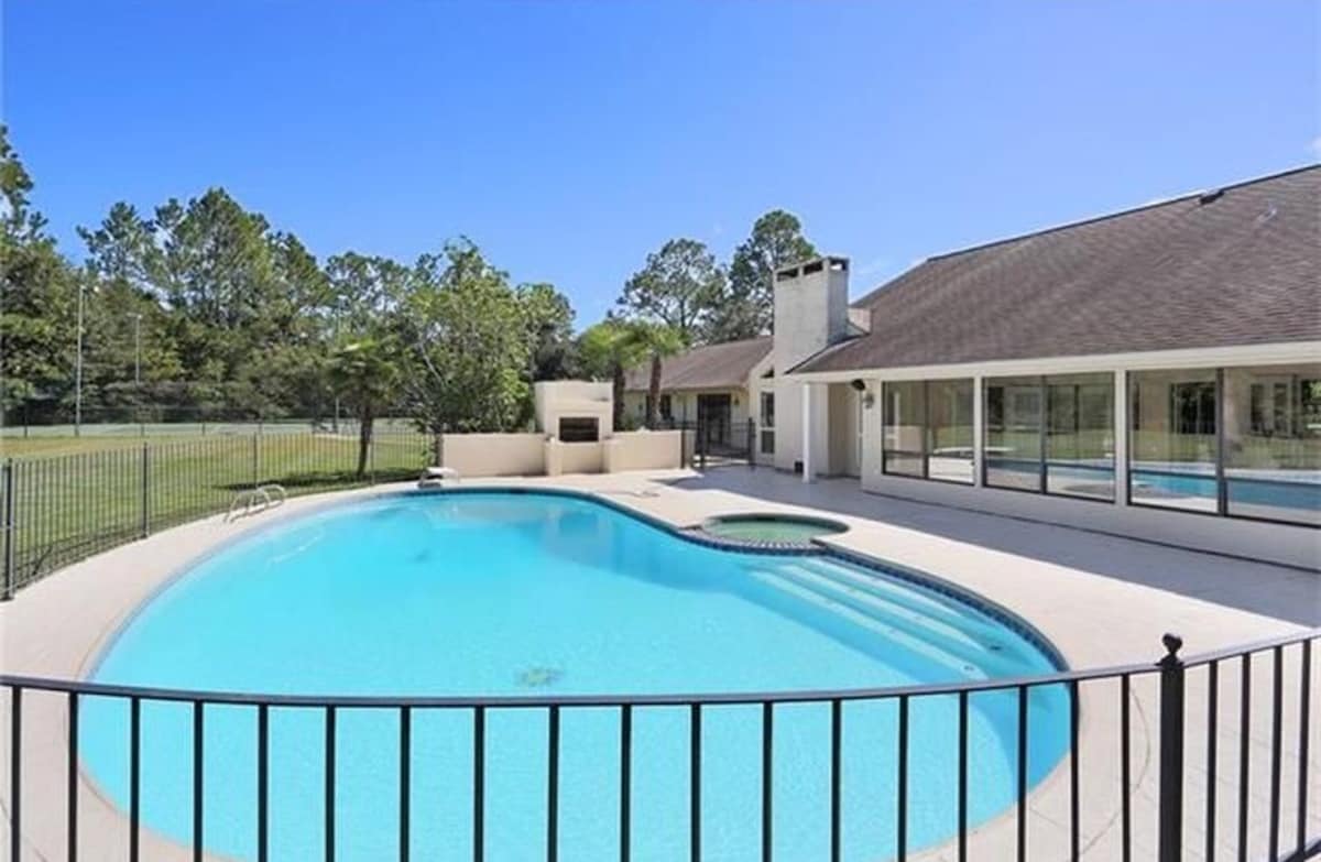A large, inviting swimming pool is surrounded by a spacious patio, featuring a jacuzzi adjacent to the pool. Tall trees provide a natural backdrop, while floor-to-ceiling windows of the nearby house enhance views of the serene outdoor space.