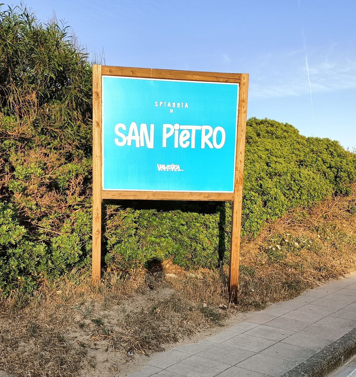 A large sign displays the name 'Spiaggia di San Pietro' in bold white letters against a bright blue background. The sign is framed by a wooden structure and surrounded by lush green foliage and sandy ground, indicating a welcoming beach area.