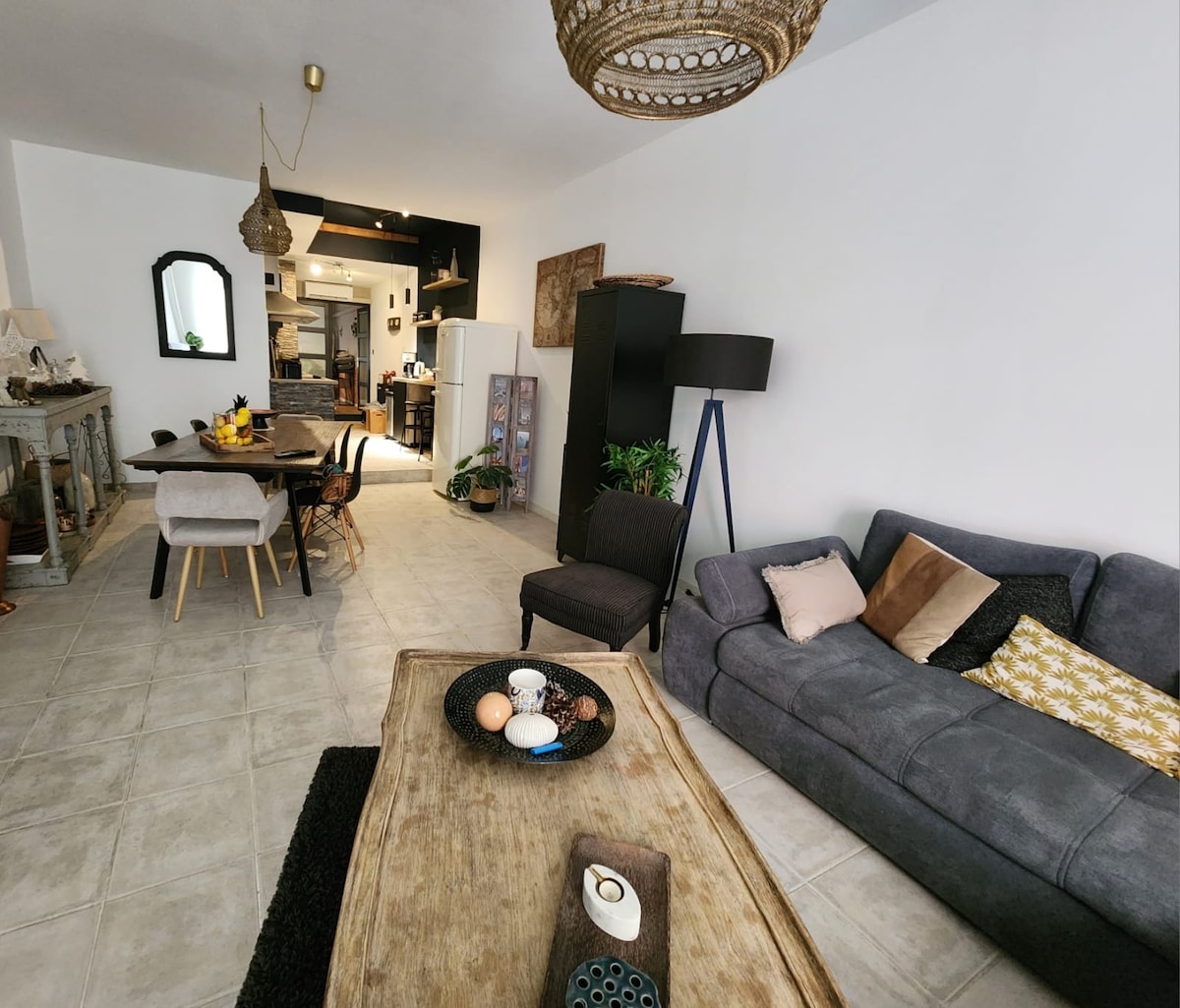 A spacious living area is displayed, featuring a comfortable sofa facing a wooden coffee table. A dining table with chairs is positioned nearby, with greenery visible in the background. The kitchen area is partially visible, showcasing modern appliances and a welcoming ambiance.