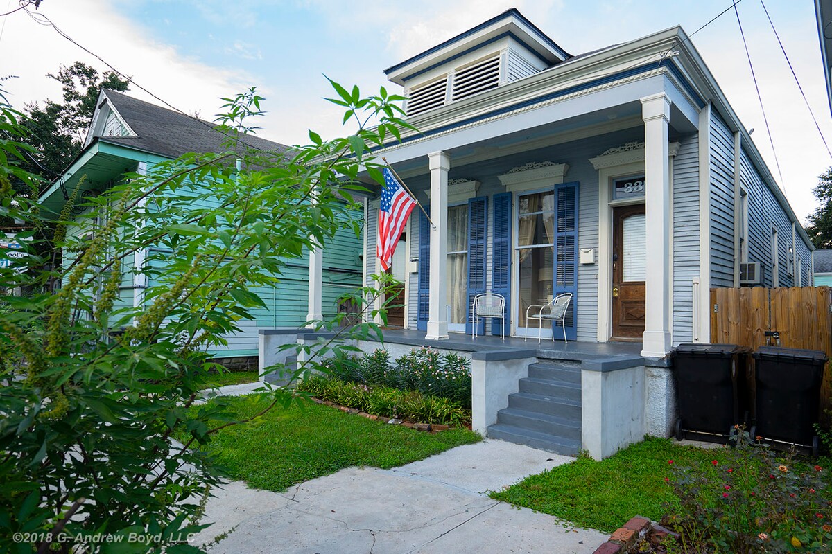 A charming exterior of a classic New Orleans home is displayed, featuring a blue and white façade. The front porch is equipped with two chairs and is adorned with an American flag. Lush greenery surrounds the entrance, contributing to the welcoming appearance.