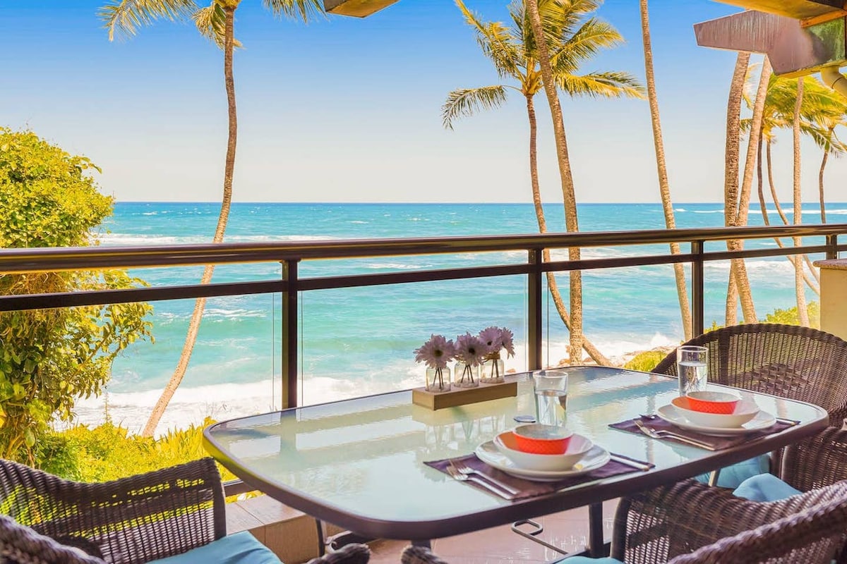 A spacious lanai features a dining table with a glass top, surrounded by dark wicker chairs. The ocean and palm trees create a stunning backdrop, with waves gently lapping at the shore in the distance.