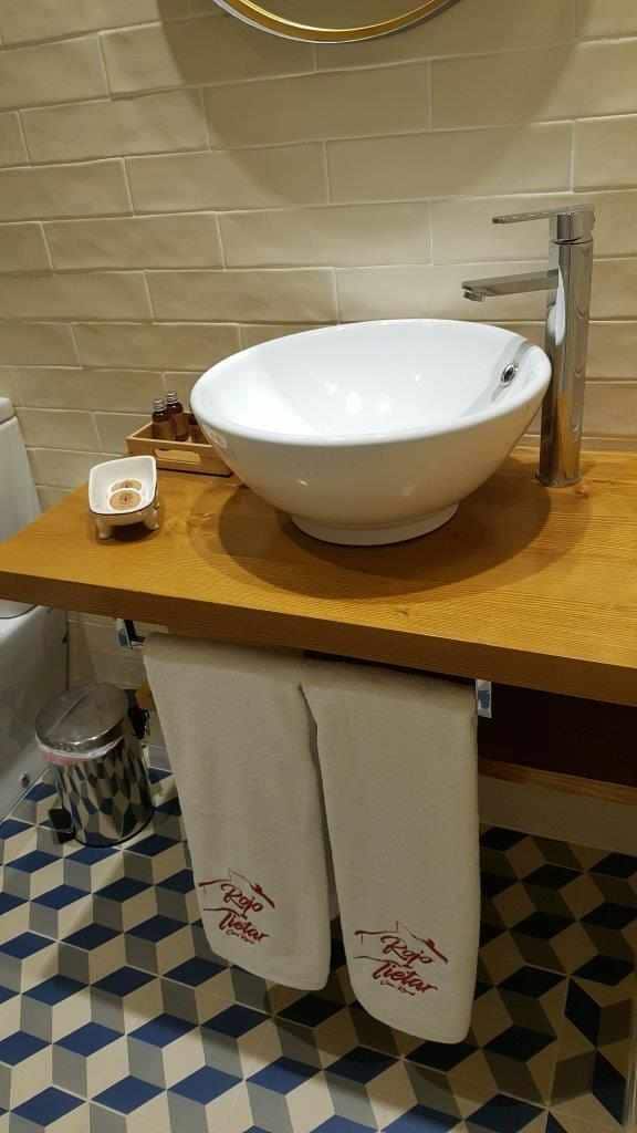 A modern bathroom sink is positioned atop a wooden counter, complemented by a sleek faucet. White towels are neatly hung nearby, featuring a subtle logo. Beneath the counter, a decorative waste bin sits among a tiled floor with a geometric pattern.