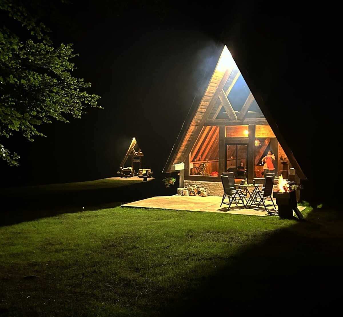 The A-frame cabin stands illuminated at night, with warm light spilling from its large windows. A spacious deck is visible, furnished with several outdoor chairs. Lush greenery surrounds the cabin, creating a serene atmosphere with a hint of smoke rising softly from a nearby fire pit.