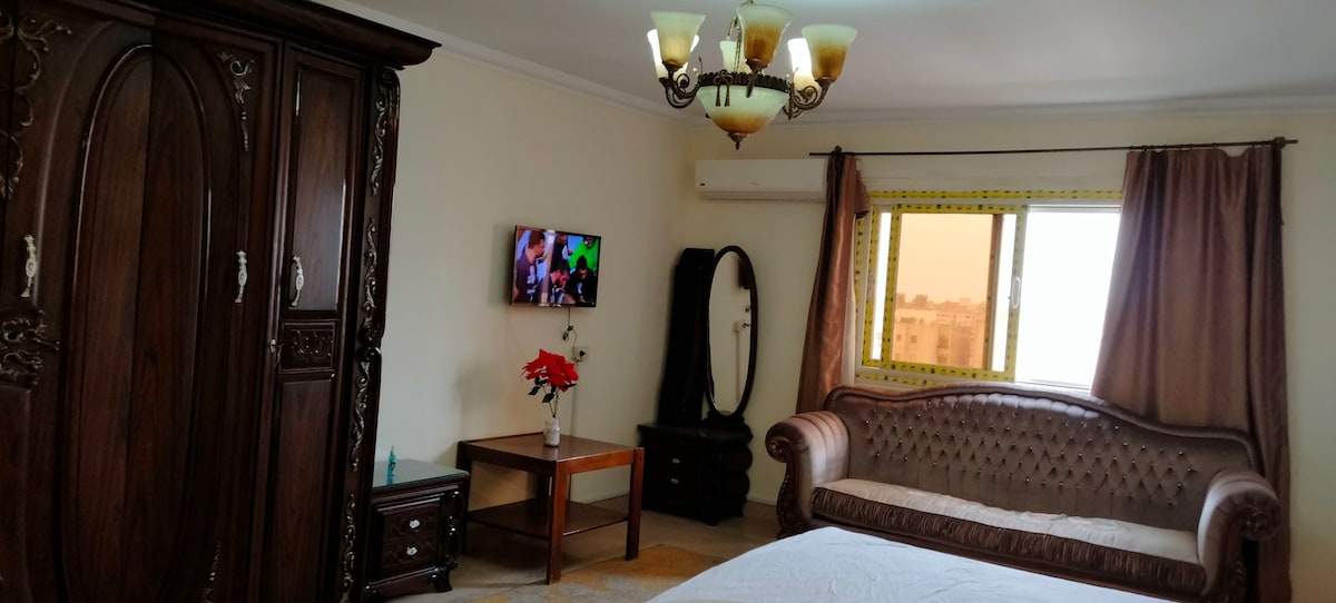 A well-furnished bedroom featuring a spacious bed and a decorative sofa. A wooden wardrobe and a table are present, along with a television mounted on the wall. Natural light enters through a large window, complementing the warm tones of the room.