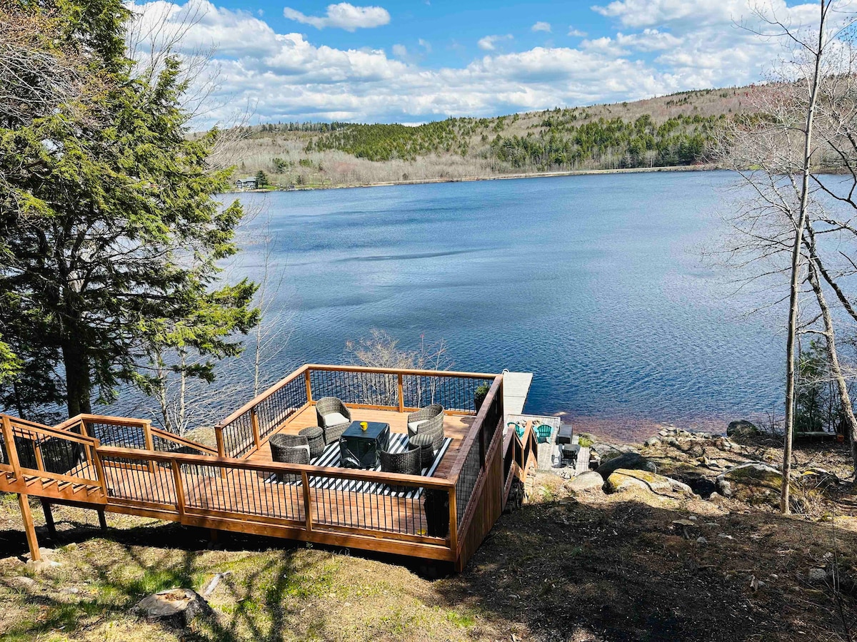 A spacious outdoor deck overlooks a serene lake, featuring a seating area with comfortable chairs and a table. The surrounding landscape includes trees and gentle slopes leading to the water's edge. Soft clouds float in a bright blue sky, enhancing the tranquil atmosphere.
