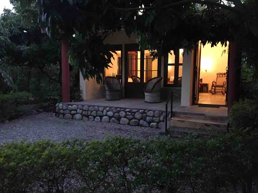 A private cottage entrance is framed by lush greenery, featuring a gravel pathway and stone steps leading to a covered porch. Two wicker chairs are positioned outside, inviting relaxation as warm light glows softly from within the cottage, illuminating the inviting space.