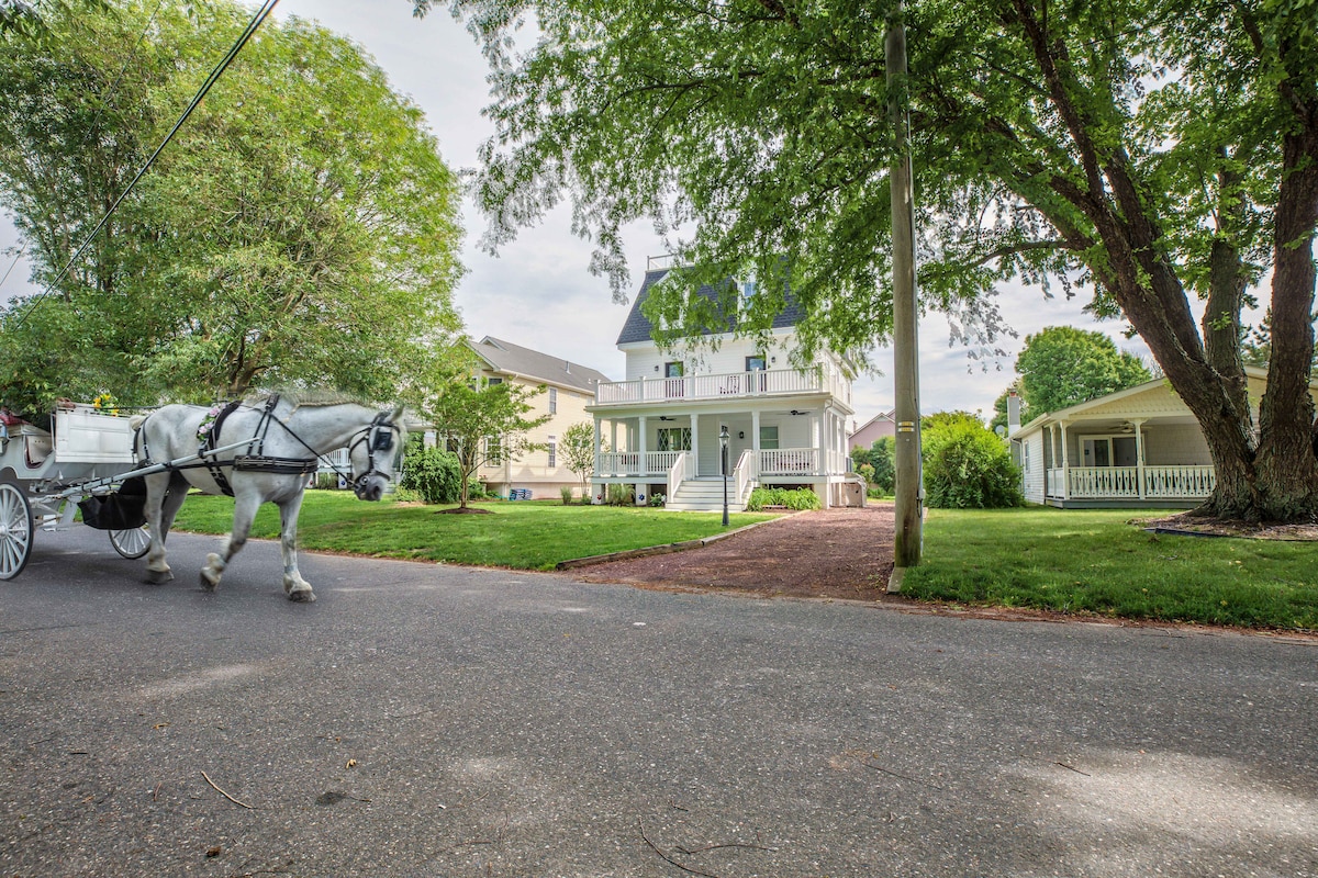 The image depicts a charming house surrounded by lush greenery and mature trees. A horse-drawn carriage is seen passing along the quiet street. The covered porch of the house invites outdoor relaxation, while the well-maintained lawn enhances the property’s appeal.