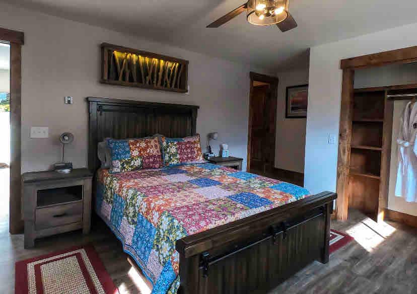 A modern bedroom is presented with a queen-sized bed featuring a colorful patchwork quilt. Bedside tables with reading lamps are positioned on either side of the bed. An oversized closet with hanging clothes is visible, along with a door leading to the outdoor space.