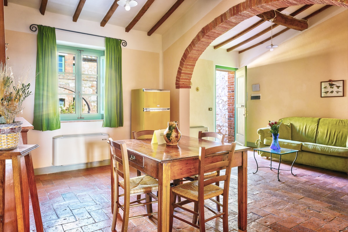 An inviting living area features exposed wooden beams overhead and terracotta flooring. A simple wooden dining table is surrounded by four chairs. The space is accented by green curtains and a cozy sofa. A vintage yellow refrigerator and a decorative plant complete the warm ambiance.