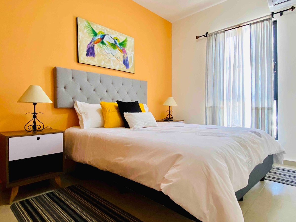 A cozy bedroom features a spacious bed with crisp white linens and decorative pillows. Bright orange walls are complemented by a colorful artwork above the headboard. Natural light enters through a large window dressed with sheer curtains, adding to the welcoming ambiance of the space.