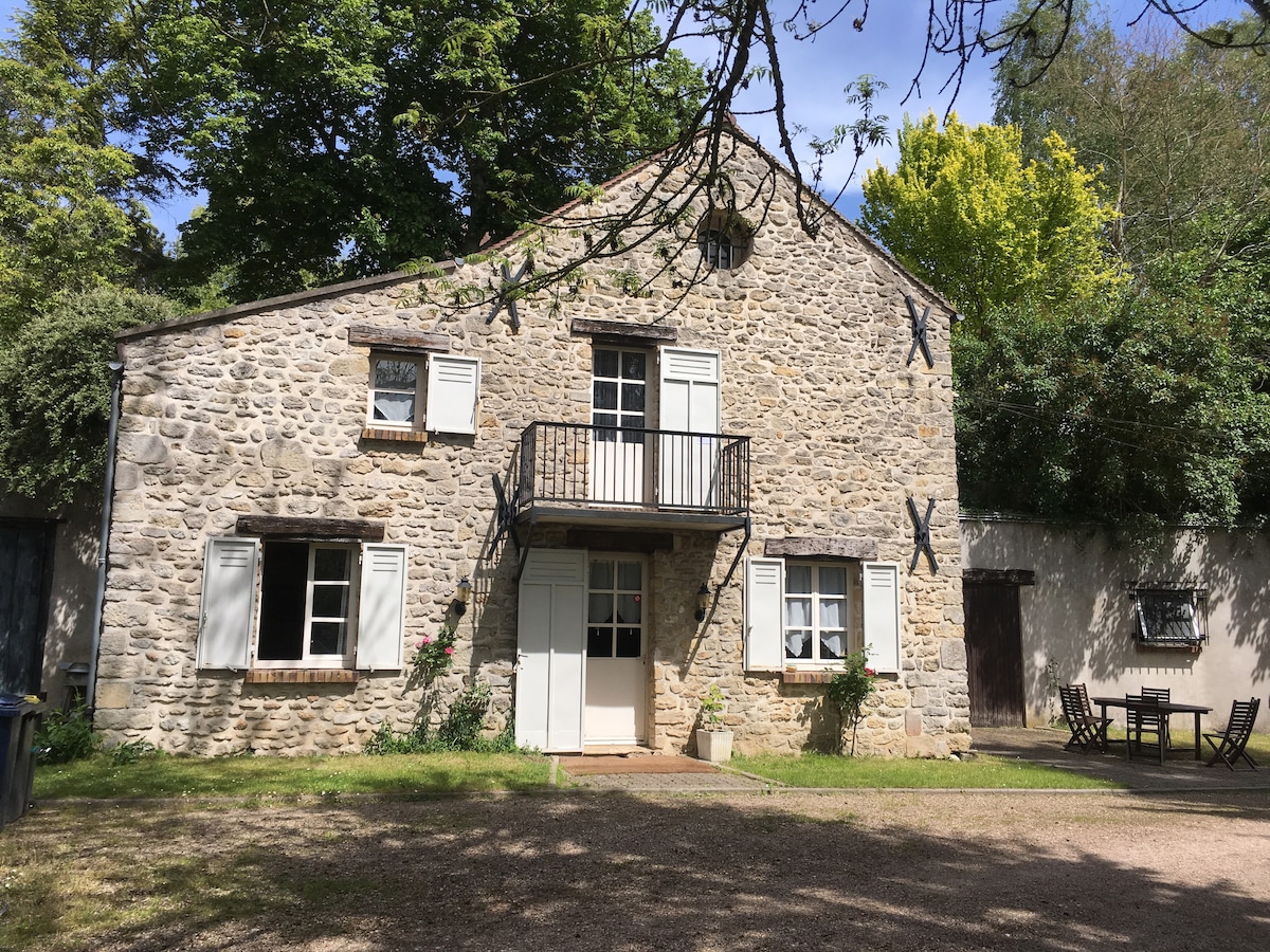 A rustic stone house is presented, featuring white shutters and a balcony. The surrounding area includes tall trees and a small outdoor seating arrangement. A gravel path leads to the front door, enhancing the home's connection to nature.