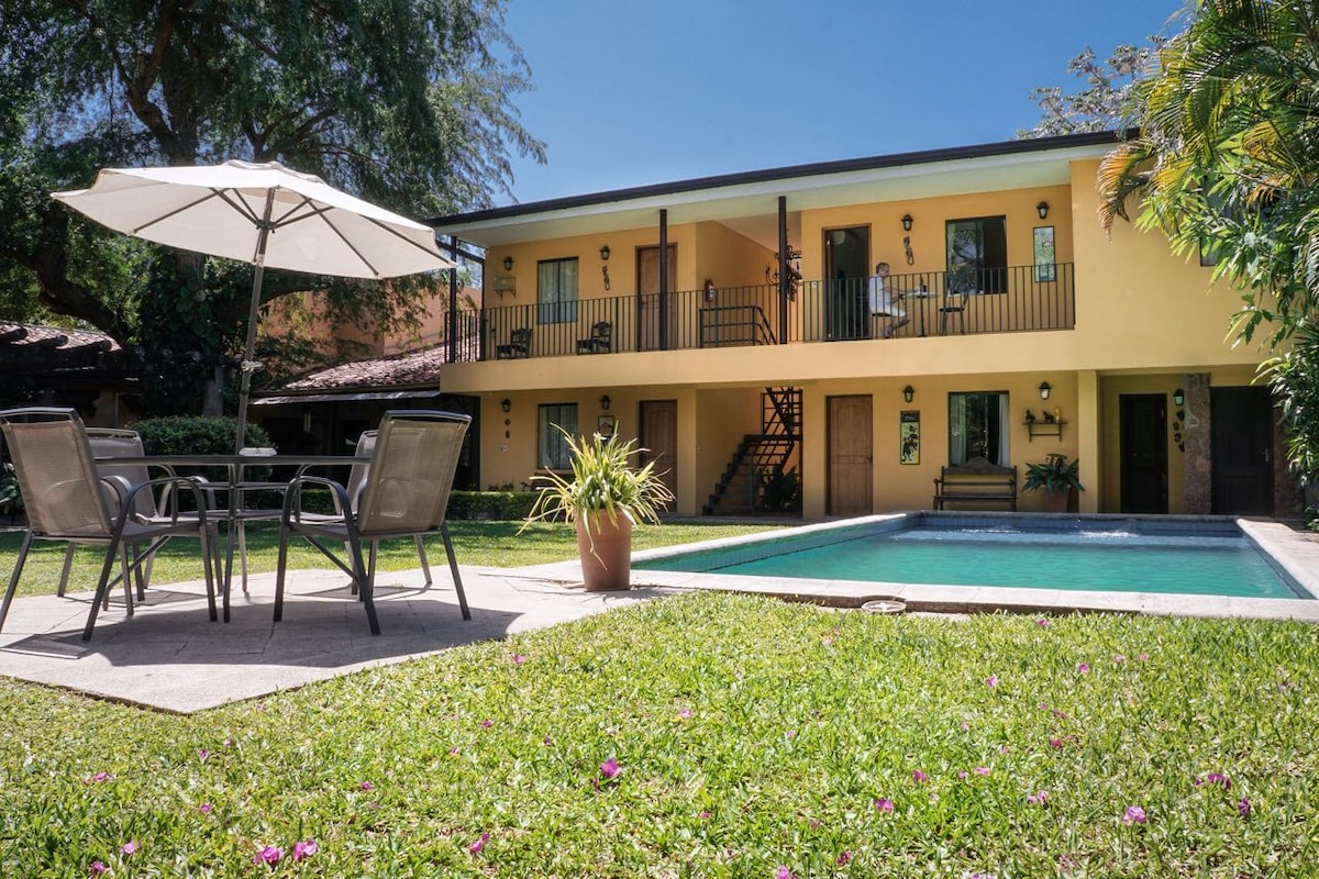 A two-story building with a warm yellow exterior is surrounded by lush green grass and colorful flowers. A patio area is furnished with a table and four chairs under a large umbrella, with a swimming pool visible in the foreground, inviting relaxation.