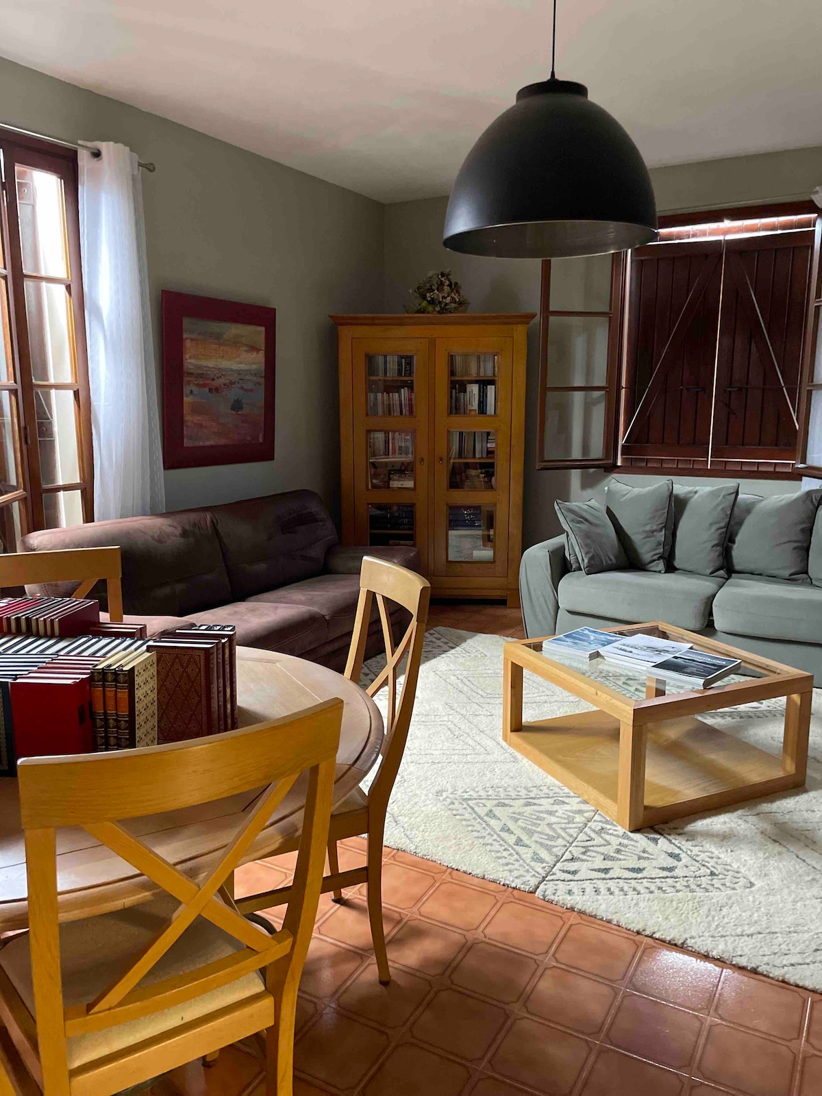 A cozy living area features a sofa set with plush cushions, complemented by a wooden coffee table at the center. Natural light fills the space through large windows, while a wooden cabinet displays books. A round dining table with wooden chairs occupies one corner.