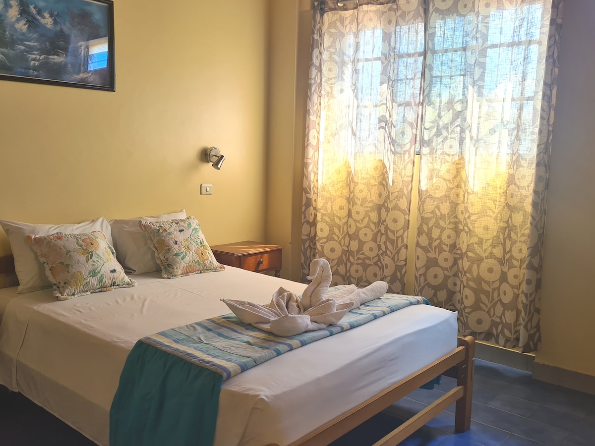 A well-lit bedroom features a spacious bed topped with decorative pillows and neatly arranged towels. Natural light filters through sheer floral curtains, illuminating the warm yellow walls. A wooden nightstand adds to the room's simple elegance, complemented by a framed artwork above the bed.