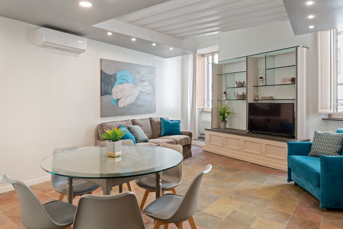 The living area is furnished with a comfortable sofa and a vibrant blue armchair, alongside a large glass dining table with multiple chairs. Natural light enters through large windows, highlighting the modern artwork displayed on the wall and the stylish shelving unit housing decorative items.