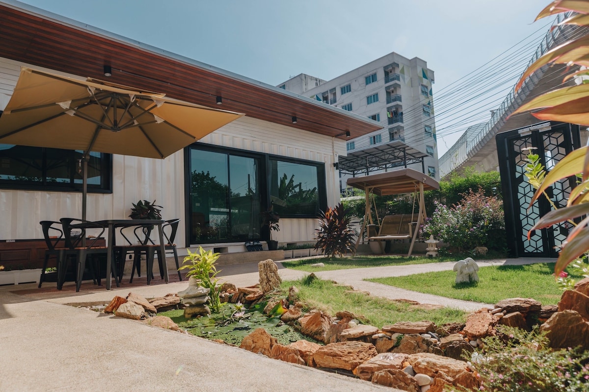 An outdoor area features a well-maintained garden with a small pond surrounded by rocks. A covered seating area with a table and four chairs is positioned next to the pond. A swing set adds a cozy touch, while large windows allow for plenty of natural light.