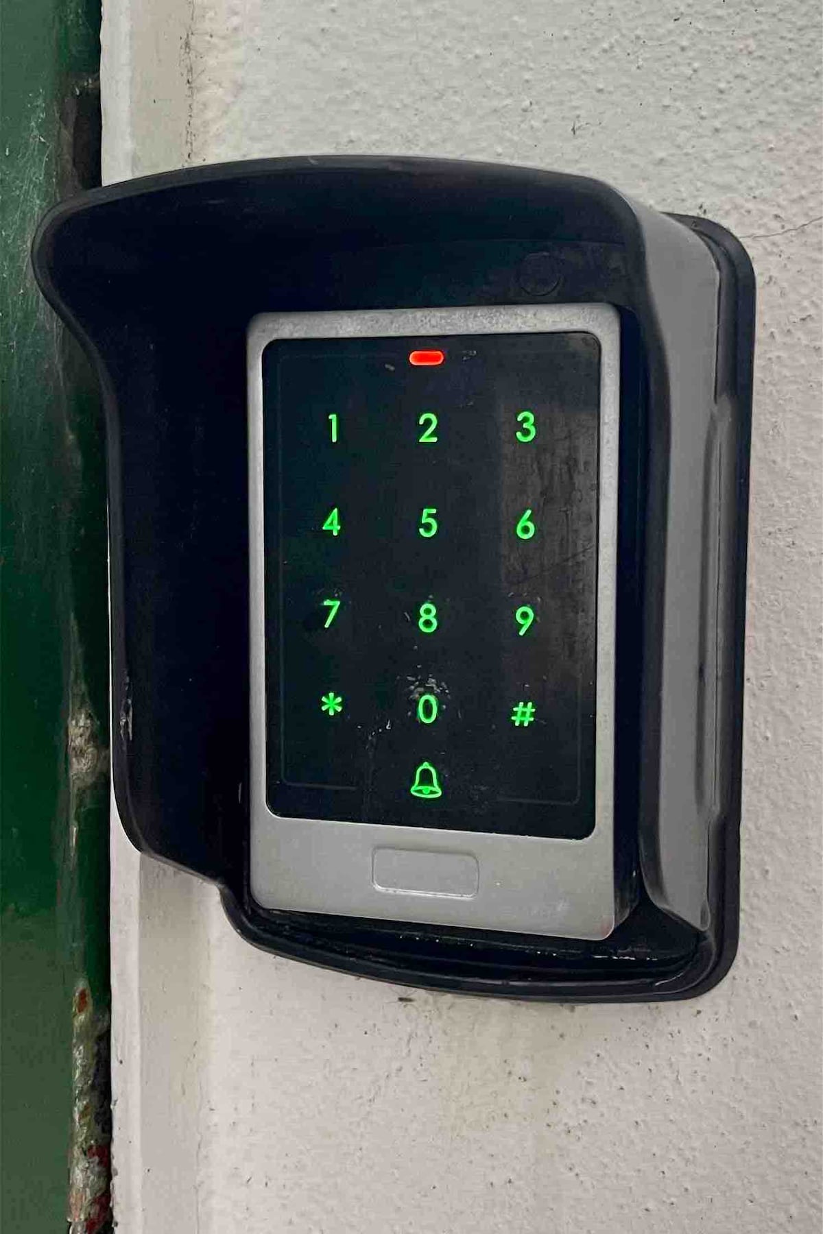 A digital keypad is mounted on a wall, featuring illuminated green numbers and symbols. A red light is visible above the screen, indicating the device's status. The surrounding area is painted in a neutral color, providing a contrast to the device.
